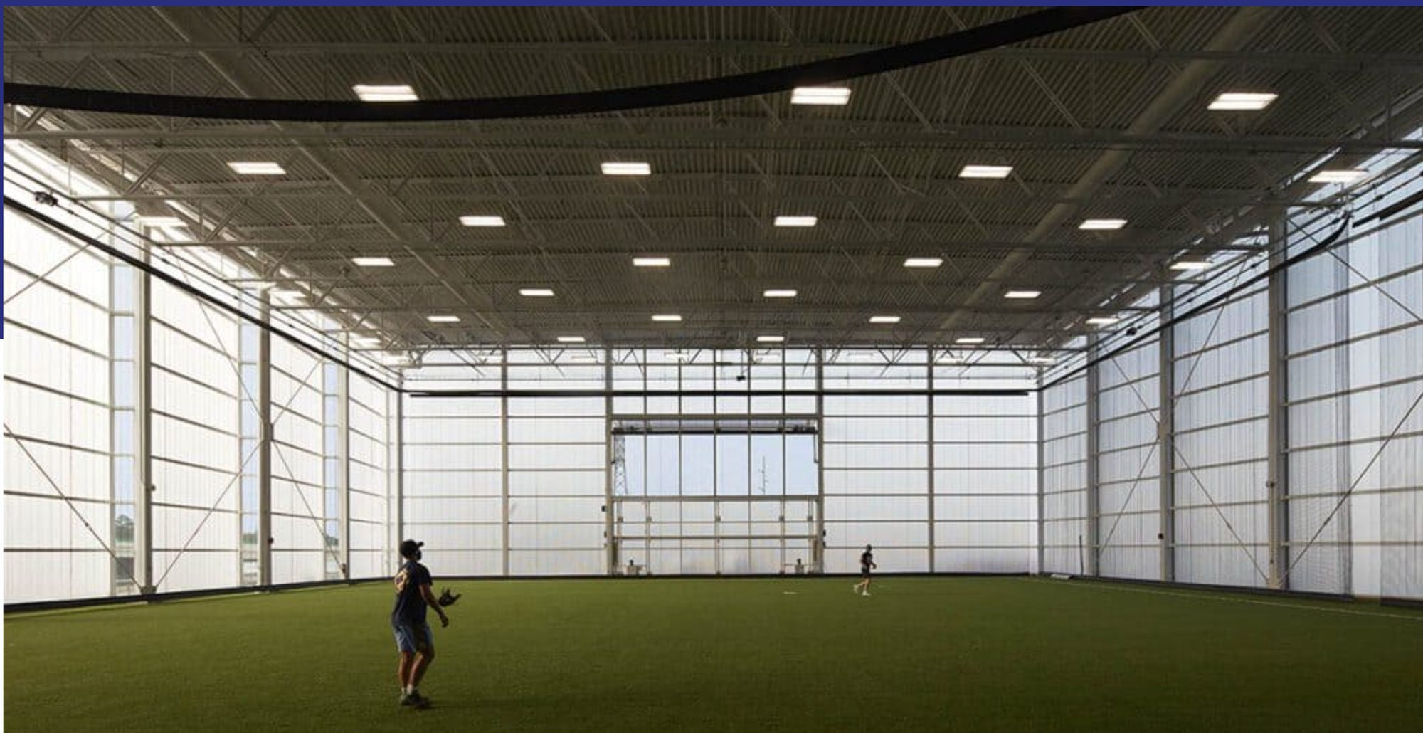 A wide-angle, interior view of a large indoor sports facility with green turf. The facility features a complex steel truss roof and three walls made of translucent, vertically ribbed polycarbonate panels, which provide diffuse natural light.